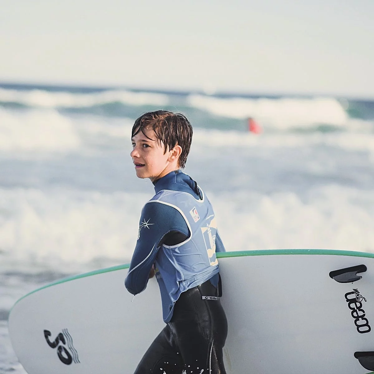 Un adolescent tient sa planche de surf sous le bras, avec l’océan en arrière-plan. Il s’apprête à faire du surf à Capbreton.