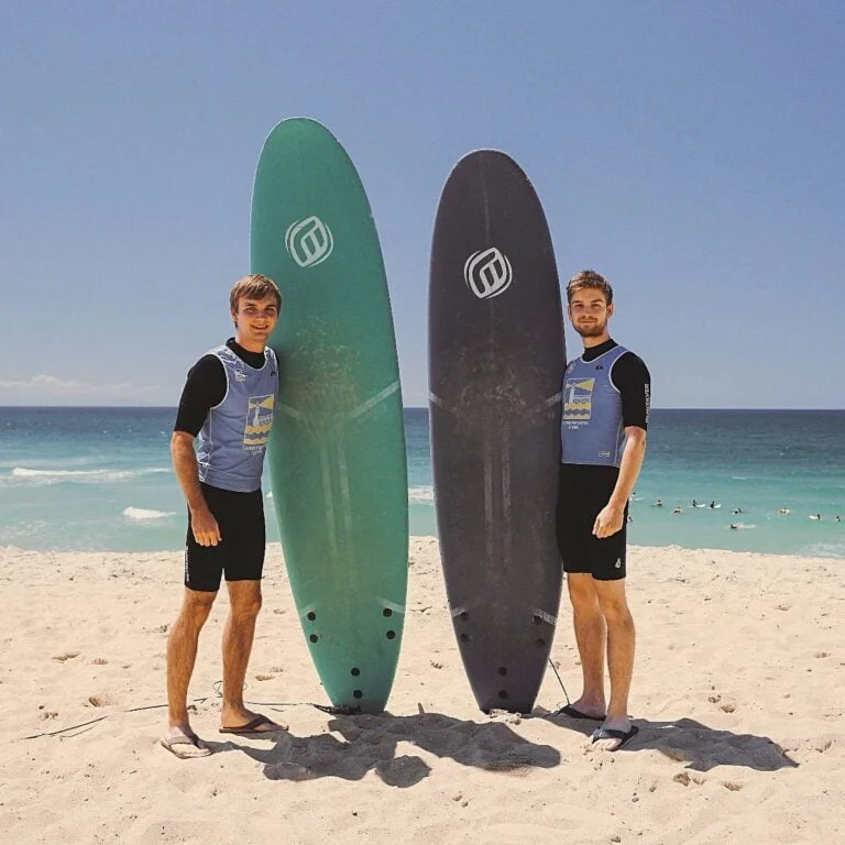 Deux jeunes hommes tiennent leurs planches de surf debout à côté d’eux sur la plage.