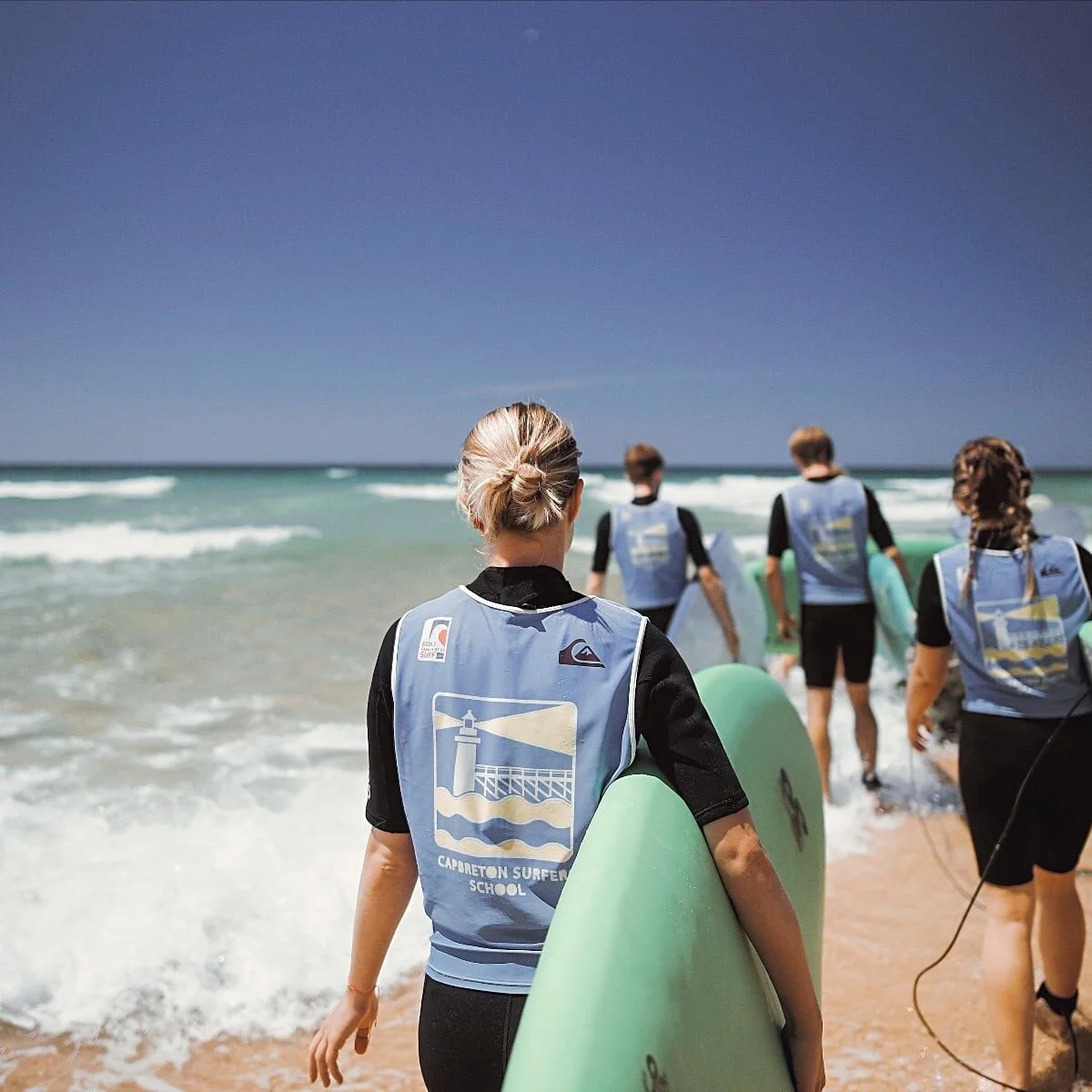 Un groupe de jeunes marche sur la plage en direction de l’océan, une planche de surf sous le bras.