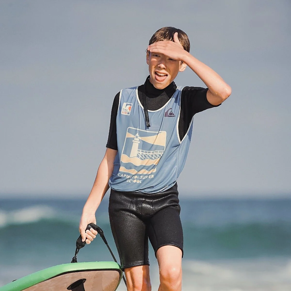 Un adolescent marche sur la plage en tenant sa planche de surf à la main tout en se protégeant les yeux du soleil.