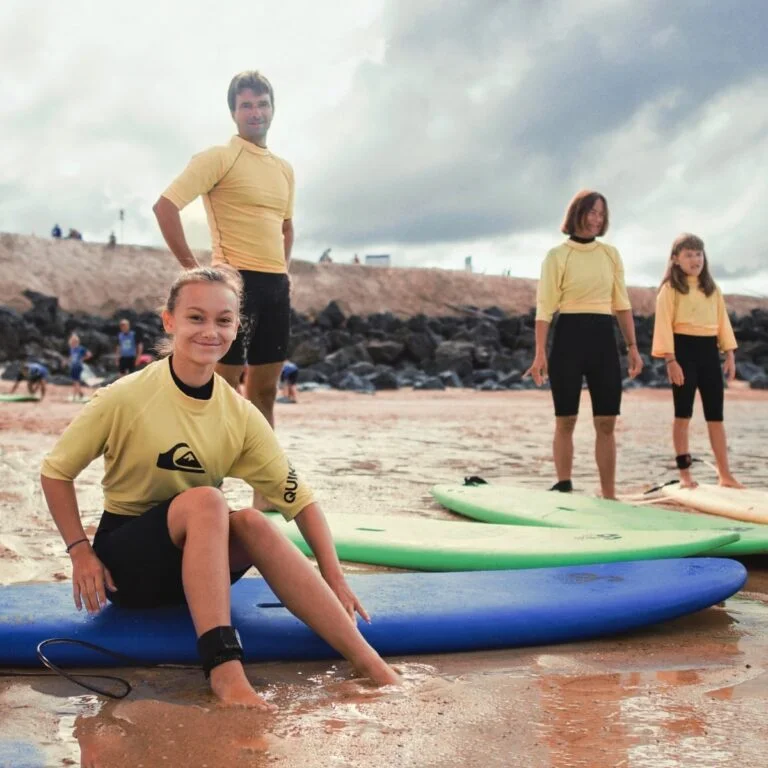 Un groupe de 4 personnes se tient sur la plage avec leurs planches de surf et sourient en regardant la caméra.