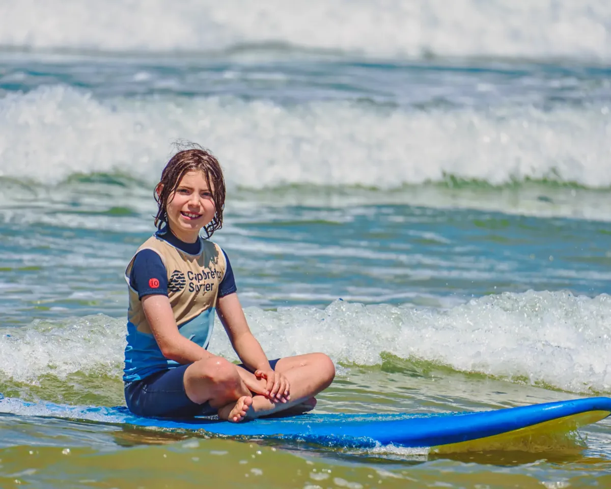 Un enfant assis sur une planche de surf bleue, souriant et regardant vers la caméra, illustrant le plaisir des cours de surf pour enfants