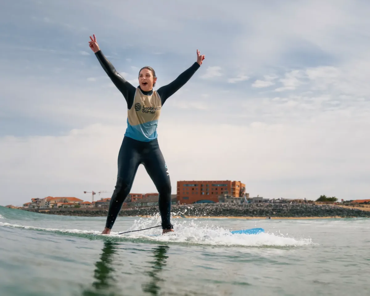 Une surfeuse souriante en combinaison noire avec un jersey aux couleurs de Capbreton Surfer, levant les bras en signe de victoire après avoir réussi à se tenir debout sur sa planche de surf