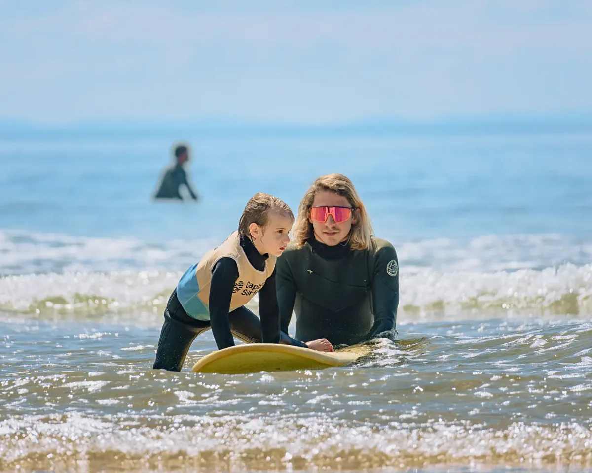 Un moniteur de surf en combinaison noire parlant à une jeune enfant et l'aide à monter sur sa planche de surf dans des eaux peu profondes