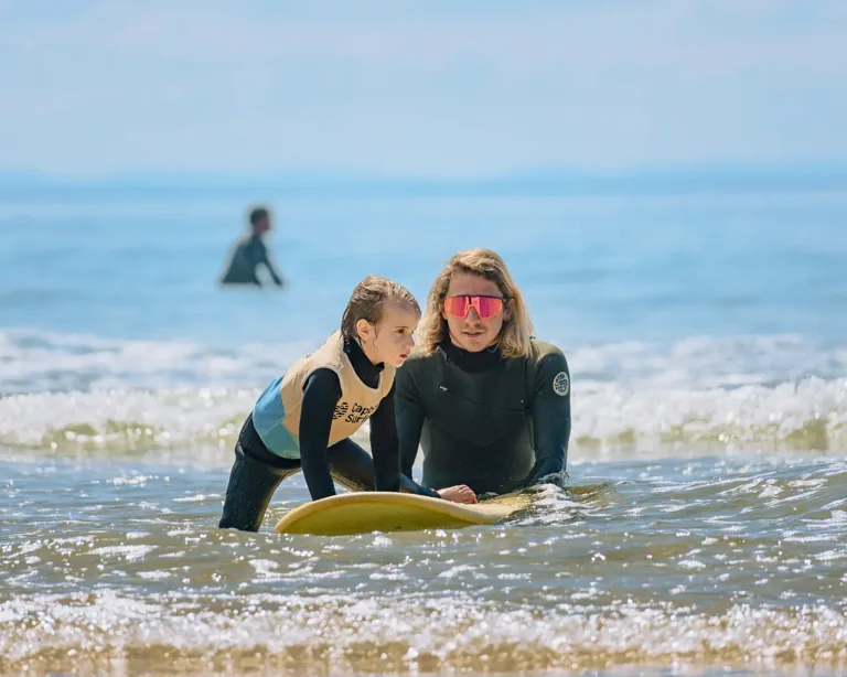 Un moniteur de surf en combinaison noire parlant à une jeune enfant et l'aide à monter sur sa planche de surf dans des eaux peu profondes