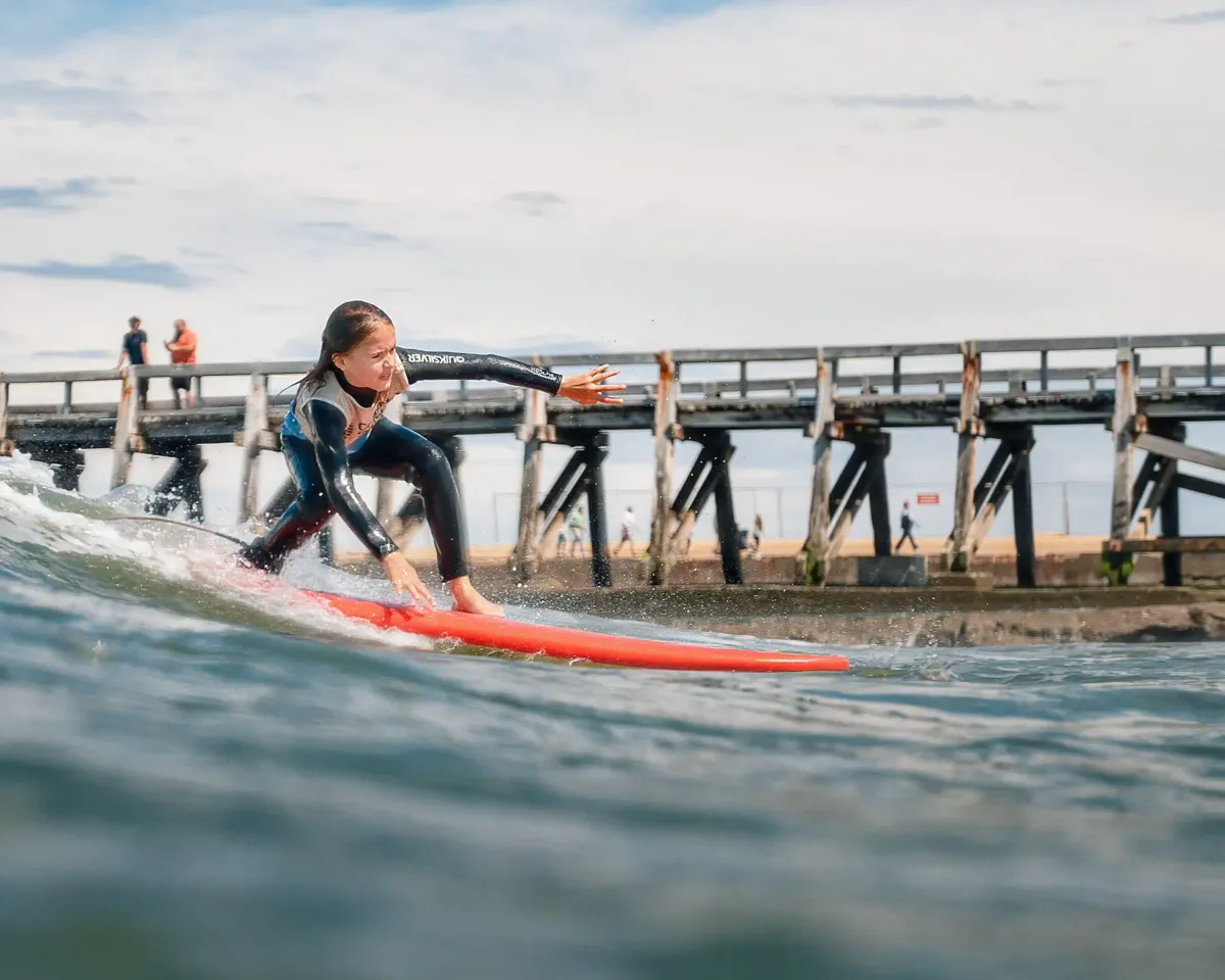 Une jeune surfeuse en combinaison noire, debout sur une planche de surf rouge, prenant une vague avec un pont en arrière-plan