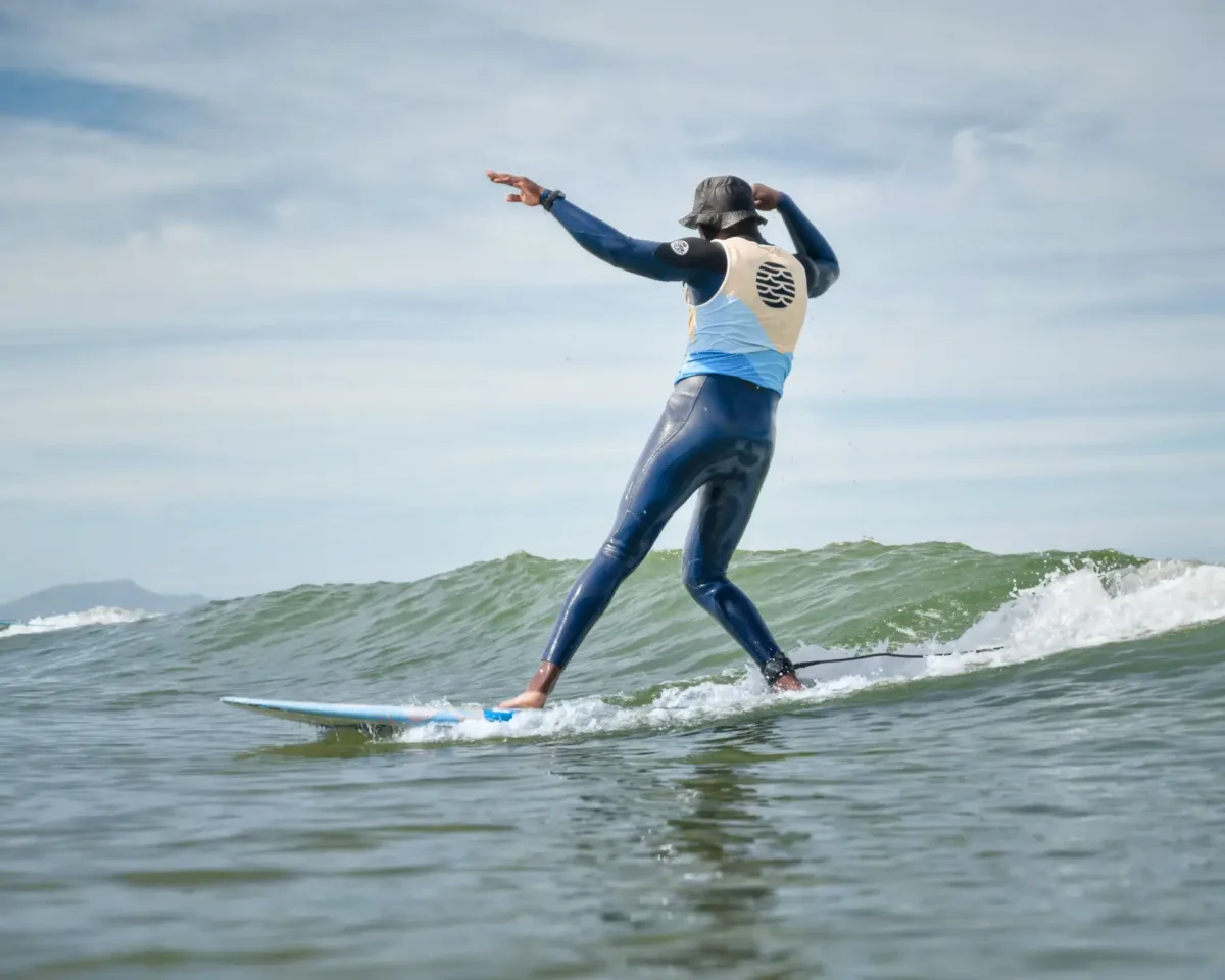 Un surfeur en combinaison, avec un jersey bleu et beige se tenant debout sur une planche de surf, comme s’il dansait sur la vague