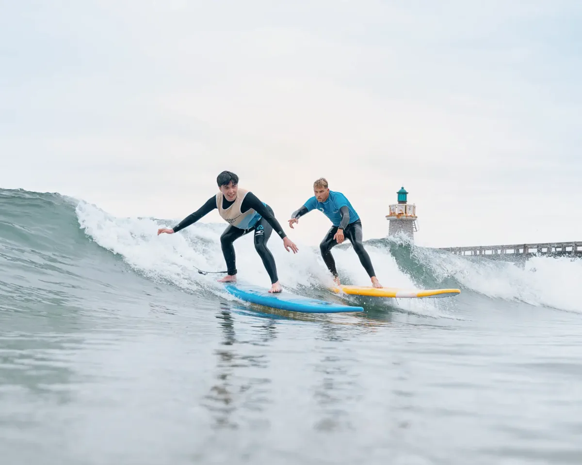 Un adolescent surfe une vague debout sur sa planche de surf bleue, tandis que le moniteur de surf, surfant lui aussi la même vague à ses côtés, lui donne des conseils sur sa posture.