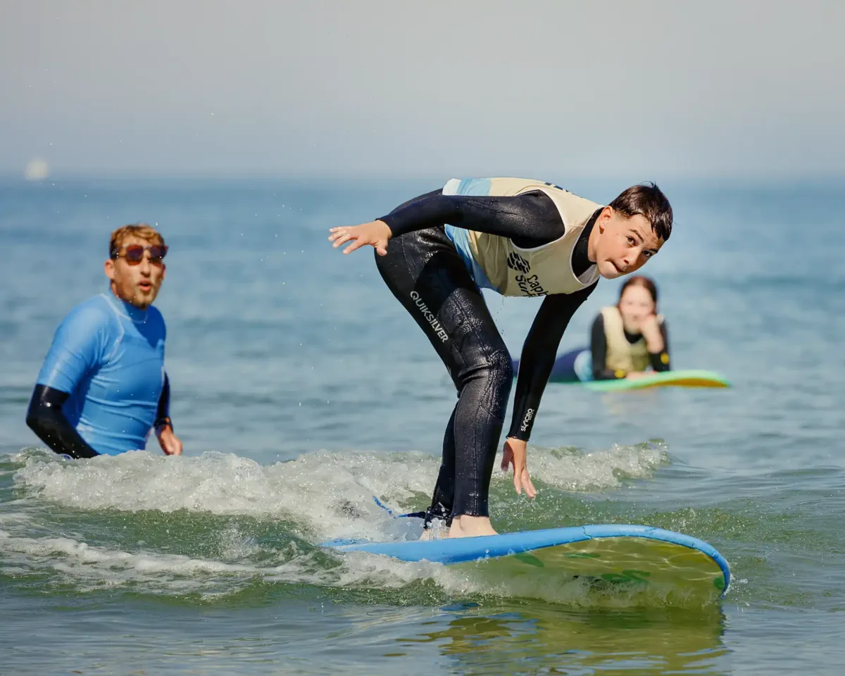 Un adolescent en combinaison de plongée surfe sur une planche de surf bleue. Il est en train de se lever sur sa planche, tandis que le moniteur de surf en arrière-plan observe sa position.
