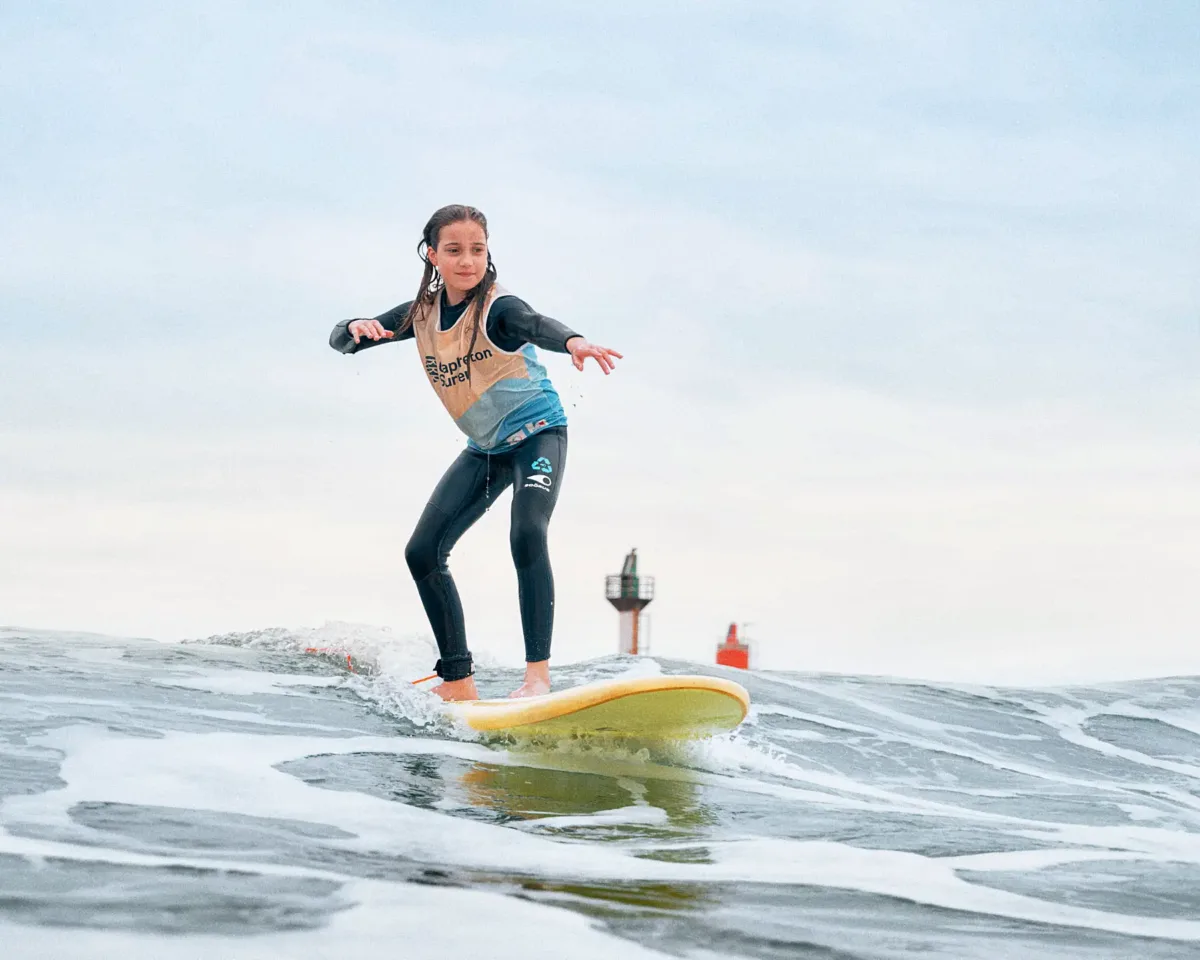 Une adolescente portant une combinaison et un lycra beige et bleu surfe sur une planche de surf jaune sur une vague calme, avec le phare de Capbreton visible au loin.