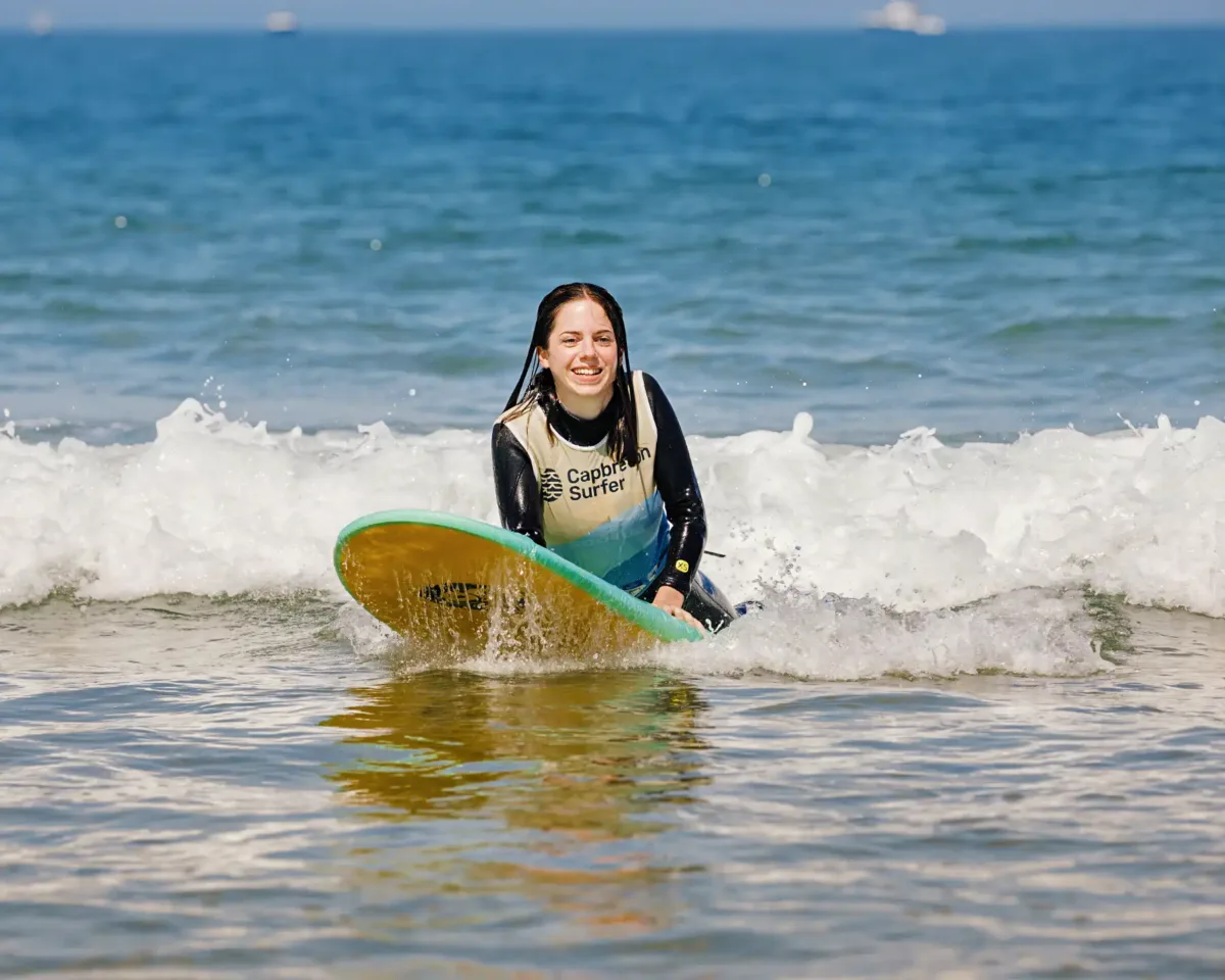 Une adolescente vêtue d'une combinaison de plongée et d'un gilet de l'école de surf est allongée sur une planche de surf dans les eaux calmes de l'océan, avec des vagues en arrière-plan. Elle sourit.