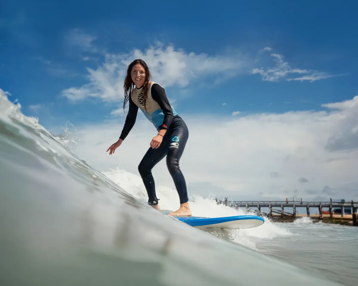 Une femme portant une combinaison de plongée surfe une vague sur une planche de surf bleue en direction de la caméra, sous un ciel partiellement nuageux.
