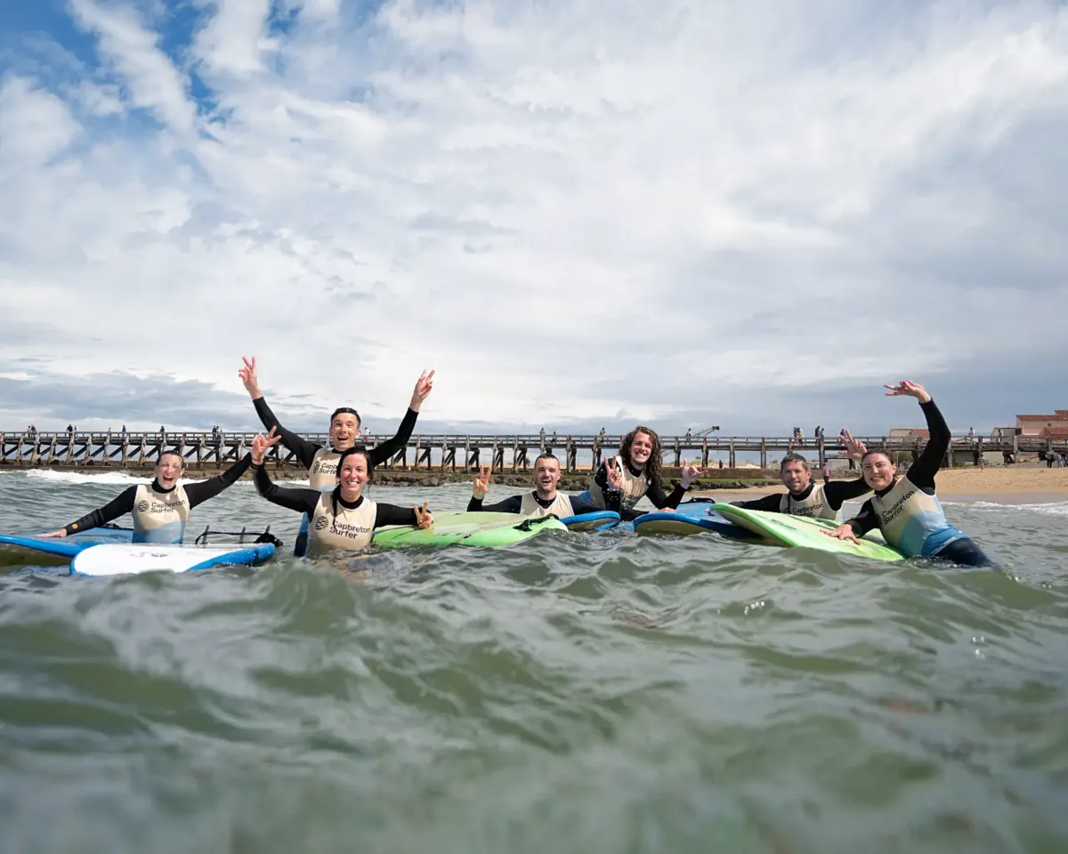 Un groupe d'adultes en combinaisons de plongée se trouve dans l'eau avec des planches de surf, souriant et levant les mains en l'air en faisant des signes de “V” avec les doigts.