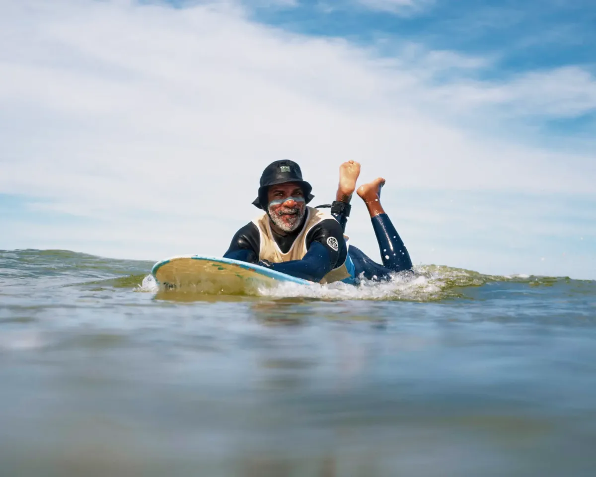 Un homme portant un chapeau noir et une combinaison de plongée est allongé sur une planche de surf dans l'océan, souriant et regardant en direction de la caméra, avec le ciel et les nuages en arrière-plan.