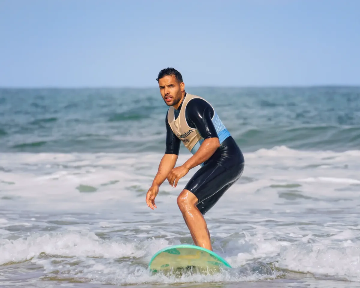 Un homme en combinaison de plongée fait face à la caméra en se tenant debout sur une planche de surf dans l'océan avec de petites vagues autour de lui.