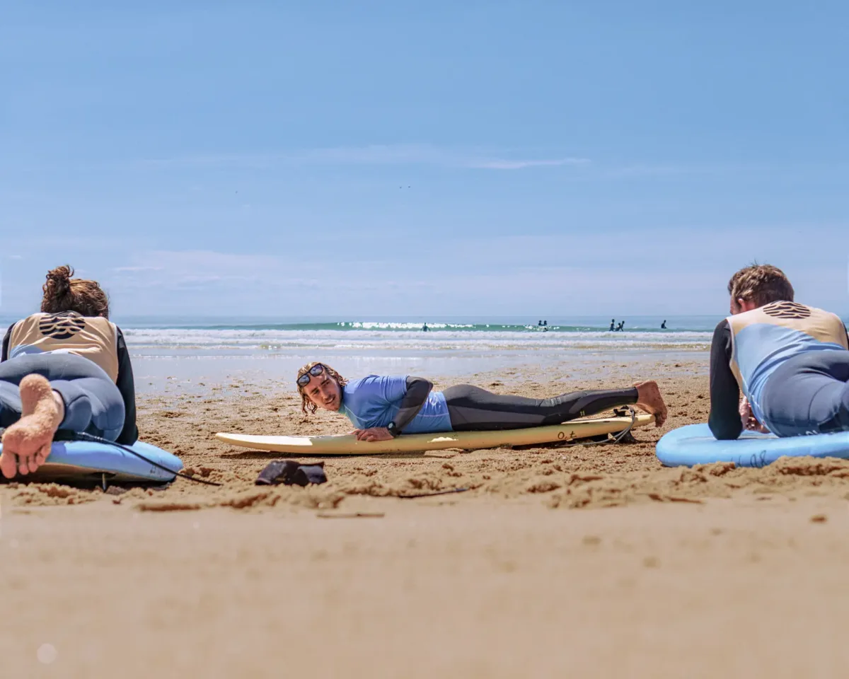 Un moniteur de surf, allongé sur sa planche sur la plage, démontre des techniques à deux adultes qui le regardent, eux aussi allongés sur leurs planches. En fond, on peut voir l'océan et les vagues au loin, sous un ciel bleu clair.