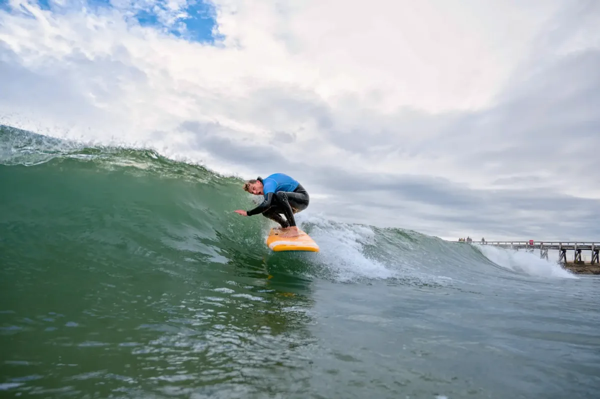 Un moniteur de surf en combinaison de plongée noir et lycra bleu surfe sur une vague qui s'apprête à se casser, en s'amusant et en prenant une pose incongrue.