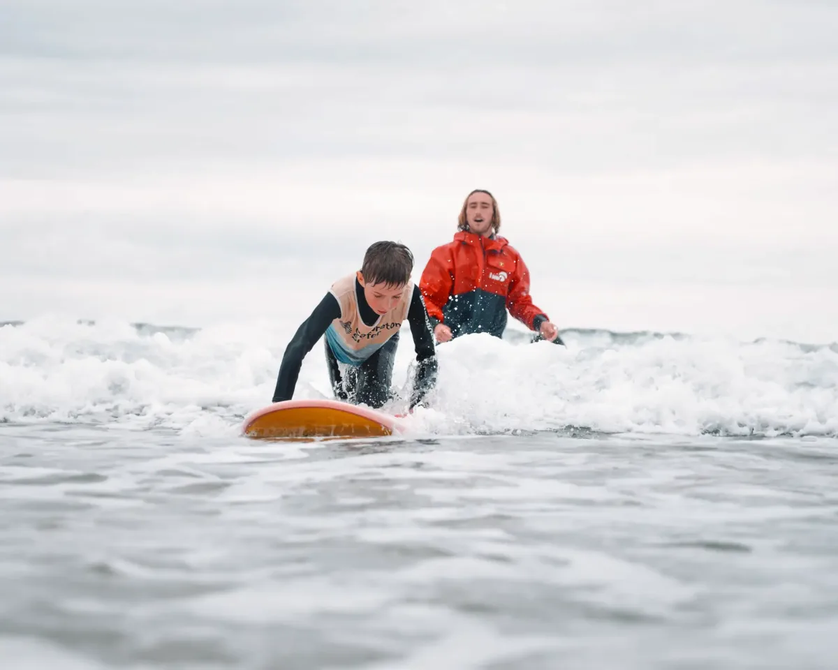 Un enfant à genoux sur sa planche de surf dans l'écume, en eaux peu profondes, sous l'œil attentif d'un moniteur en parka rouge