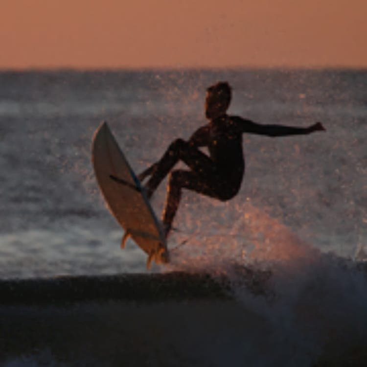 Un jeune homme en surf saute par-dessus une vague au coucher du soleil. Sa silhouette se détache en contrejour devant la mer.